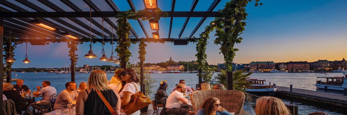 1200x400_category-pages-2026-03-28b People dining at a waterfront restaurant in Stockholm on Strandvägen during sunset with infrared heaters and scenic harbor view