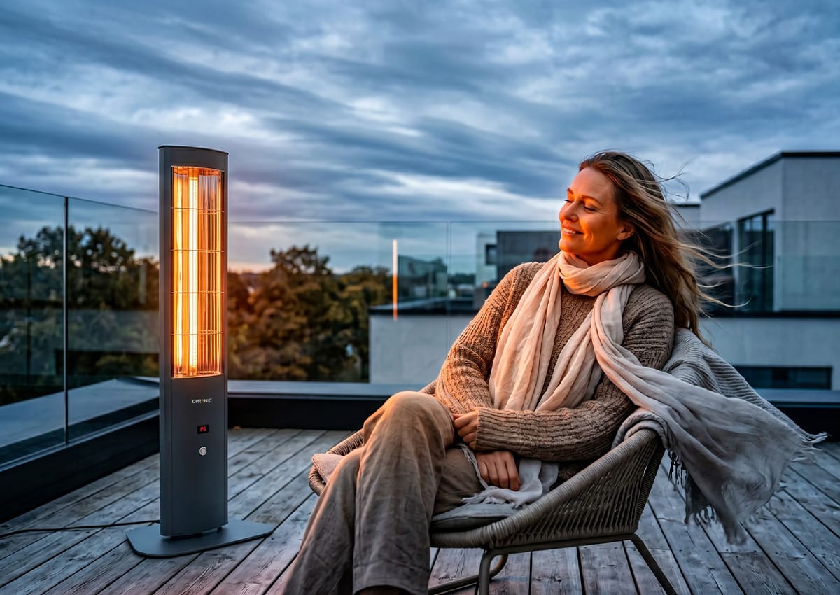 Kvinna sitter avslappnad på blåsig taketerrass i höstvind, värmad av en Opranic THOR S70R infraröd golvvärmare med glödande orange sken, infraröd värme som fungerar trots vind och kyla.